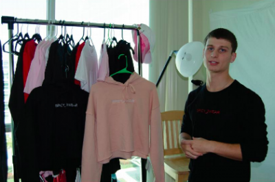 Person standing next to a rack of hoodies in a room with a chair and window.